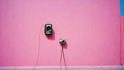 The primary subject of the image is a pair of old-fashioned cameras, which are placed on a pink wall. The setting is a room with a pink wall, and the visual style appears to be a photo. The colors and mood of the image are predominantly pink, giving it a warm and nostalgic atmosphere.