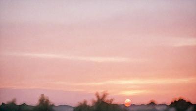 The primary subject of the image is a beautiful sunset, with the sun setting behind a hill. The setting is a cloudy sky, and the visual style is a photo. The colors and mood of the image are warm and serene, with the sun's rays creating a glowing effect on the clouds and the hill.