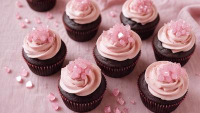 The image features a table with a dozen cupcakes, each topped with a pink sugar rose. The cupcakes are arranged in a visually appealing manner, creating an enticing display. The table is the primary subject of the image, and the cupcakes are the main objects, scene, or focus of the image. The setting is a dining table, and the visual style is a photo. The colors and mood of the image are predominantly pink, which adds a sense of warmth and sweet