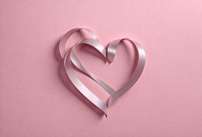 The primary subject of the image is a heart-shaped piece of ribbon. The heart is made of two pieces of ribbon, one on top of the other, and is placed on a pink background. The visual style of the image is a close-up of the heart-shaped ribbon, showcasing its details. The colors and mood of the image are predominantly pink, giving it a warm and romantic atmosphere.