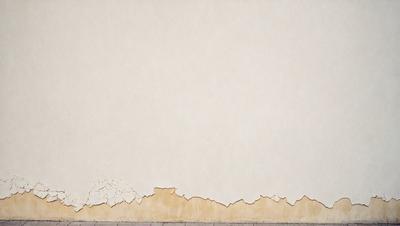 The primary subject of the image is a white wall with a peeling paint surface. The setting is a room with a white wall, and the visual style is a photo. The dominant colors are white and brown, and the overall atmosphere is somewhat aged or worn.