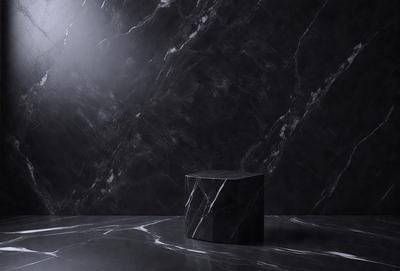 The primary subject of the image is a black marble block or a black cube, which is placed on a black marble countertop. The setting is a dark room with a black marble wall, creating a dramatic and moody atmosphere. The visual style is a close-up of the black marble block, emphasizing its texture and details. The colors and mood of the image are predominantly black, giving it a sleek and modern appearance.