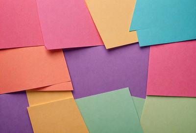 The primary subject of the image is a collection of colorful paper, including pink, yellow, green, and blue. These papers are arranged in a visually appealing manner, creating a vibrant and lively scene. The setting is a table, and the visual style is a close-up of the colorful papers. The dominant colors and overall atmosphere of the image are cheerful, lively, and full of energy.