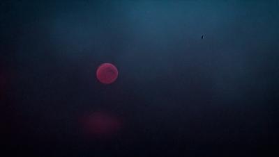 The primary subject of the image is a red moon, which is the main focus of the scene. The setting is a dark, cloudy sky, creating a moody atmosphere. The visual style is a photo, capturing the moon's glow against the night sky. The colors and mood of the image are dominated by the red hue of the moon and the darkness of the sky, evoking a sense of mystery and wonder.