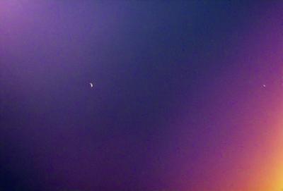 The primary subject of the image is the moon, which is visible in the sky. The setting is a dark, cloudy night with a purple hue. The visual style is a photo, capturing the moon in its natural environment. The colors and mood of the image are predominantly purple, creating a serene and atmospheric scene.