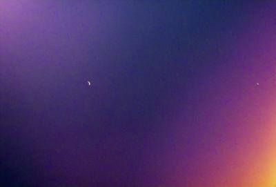 The primary subject of the image is a crescent moon, which is visible in the sky. The setting is a dark, cloudy night with a purple hue. The visual style is a photo, capturing the moon in its natural environment. The colors and mood of the image are predominantly purple, creating a serene and atmospheric scene.