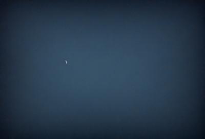 The primary subject of the image is a moon, which is visible in the sky. The setting is a dark, cloudy night with a blue sky. The visual style is a photo, capturing the moon and the night sky. The colors and mood of the image are predominantly dark and moody, with the moon being the main source of light in the scene.
