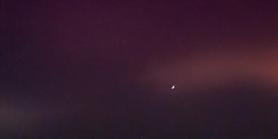 The primary subject of the image is the moon, which is prominently visible in the sky. The setting is a dark, cloudy night with a purple hue, creating a moody atmosphere. The visual style is a photo, capturing the moon's silhouette against the night sky. The colors and mood of the image are predominantly dark and dramatic, with the moon serving as the focal point against the backdrop of the cloudy sky.