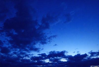 The primary subject of the image is a cloudy blue sky with a few clouds scattered throughout. The setting is a nighttime sky, with the sun either setting or rising, creating a serene atmosphere. The visual style is a natural, outdoor scene, with no indication of digital art or illustration. The colors and mood are predominantly blue, with some white clouds, creating a calm and peaceful ambiance.