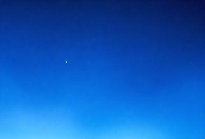 The primary subject of the image is the moon, which is visible in the sky. The setting is a clear blue sky, with no clouds or other obstructions. The visual style is a photo, capturing the moon and the sky in a natural and unaltered manner. The colors and mood of the image are predominantly blue, creating a serene and peaceful atmosphere.