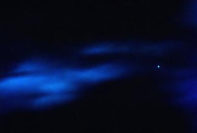 The primary subject of the image is a blue cloud in the sky. The setting is a dark night, with the cloud being the main focus of the scene. The visual style appears to be a photo, capturing the cloud's vibrant blue hue. The colors and mood of the image are predominantly dark and moody, with the blue cloud standing out against the night sky.