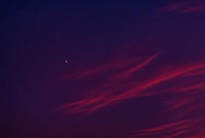 The primary subject of the image is a moon, which is visible in the sky. The setting is a dark, cloudy night with a purple hue. The visual style is a photo, capturing the moon and its surroundings. The colors and mood of the image are predominantly purple, creating a serene and atmospheric scene.