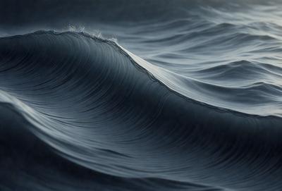 The primary subject of the image is a large wave in the ocean. The wave is the main focus of the image, and it is captured in a close-up shot. The setting is the ocean, with the wave being the primary background. The visual style of the image is a photo, capturing the natural beauty of the ocean and the wave. The colors and mood of the image are predominantly blue, with the wave appearing dark and powerful.