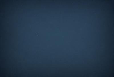 The primary subject of the image is the moon, which is visible in the sky. The setting is a dark, cloudy night with a blue sky. The visual style is a photo, capturing the moon and the night sky. The colors and mood of the image are predominantly dark and moody, with the moon being the main source of light in the scene.