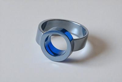 The primary subject of the image is a silver ring with a blue circle in the center. The ring is placed on a white surface, which serves as the setting or background. The visual style of the image is a close-up of the ring, showcasing its details. The colors and mood of the image are predominantly white and silver, with the blue circle adding a touch of color and contrast.