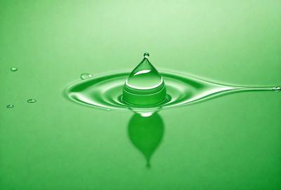 The primary subject of the image is a single drop of water, which is captured in a close-up shot. The setting is a green surface, possibly a wall or a table, which provides a contrasting background for the water drop. The visual style of the image is a photo, showcasing the water drop in its natural form. The colors and mood of the image are predominantly green, giving it a calm and serene atmosphere.