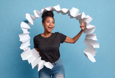 The primary subject of the image is a woman wearing a black shirt and jeans. She is pointing at a circle of paper, which is the main object in the scene. The setting is a blue background, and the visual style appears to be a photo. The woman's expression and body language convey a sense of excitement and engagement with the paper circle.