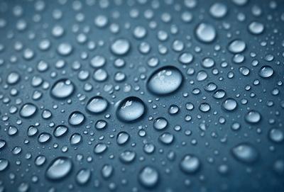 The primary subject of the image is a close-up of a blue surface with water droplets on it. The setting is a dark background, which contrasts with the blue surface. The visual style is a photo, capturing the details of the water droplets on the surface. The colors and mood of the image are predominantly dark and moody, with the blue surface and water droplets creating a sense of depth and intrigue.