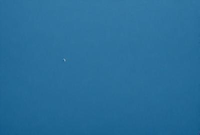 The primary subject of the image is the moon, which is visible in the sky. The setting is a clear blue sky, with no clouds or other obstructions. The visual style is a photo, capturing the moon in its natural environment. The colors and mood of the image are predominantly blue, creating a serene and peaceful atmosphere.