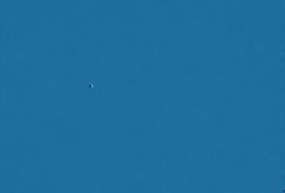 The primary subject of the image is a single, small, white dot in the middle of a clear blue sky. The setting is a vast, open space with no visible landmarks or objects. The visual style is a simple, clean, and minimalist representation of the sky and the dot. The colors and mood are predominantly blue, with a sense of calmness and tranquility.