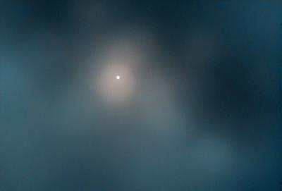 The primary subject of the image is a bright moon, which is the main focus of the scene. The moon is located in the middle of the image, surrounded by a cloudy sky. The setting appears to be a dark, cloudy night, with the moon being the only source of light in the scene. The visual style of the image is a photo, capturing the beauty of the moon in the cloudy sky. The colors and mood of the image are predominantly dark and moody, with the moon being the only bright