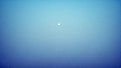 The primary subject of the image is the moon, which is seen in the sky above the Earth. The setting is a vast, open space with a blue sky, and the visual style is a combination of a photo and a drawing. The colors and mood of the image are predominantly blue, creating a serene and peaceful atmosphere.