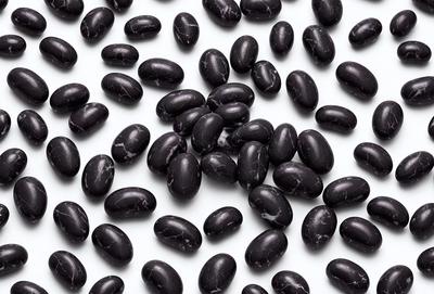 The primary subject of the image is a large group of black beans. They are arranged in a pattern on a white surface, creating a visually striking contrast. The setting is a white background, which allows the beans to stand out as the main focus of the image. The visual style is a close-up of the beans, showcasing their texture and details. The colors and mood of the image are predominantly black and white, giving it a clean and minimalist appearance.