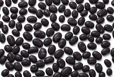The primary subject of the image is a large collection of black beans. The beans are arranged in a pattern, covering the entire image. The setting is a white background, which provides a clean and uncluttered appearance. The visual style is a close-up of the beans, showcasing their details and texture. The colors and mood of the image are predominantly black, giving it a bold and striking appearance.