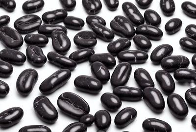 The primary subject of the image is a large collection of black beans. The beans are arranged in a pattern, covering the entire surface of the image. The setting is a white background, which provides a clean and uncluttered appearance. The visual style is a close-up of the beans, showcasing their details and texture. The colors and mood of the image are predominantly black, giving it a bold and striking appearance.
