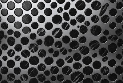 The primary subject of the image is a close-up of a patterned surface, which appears to be a metal or stainless steel material. The setting is a black background, which contrasts with the patterned surface. The visual style is a close-up of the pattern, which showcases the intricate details of the design. The colors and mood of the image are predominantly black and white, giving it a modern and sleek appearance.