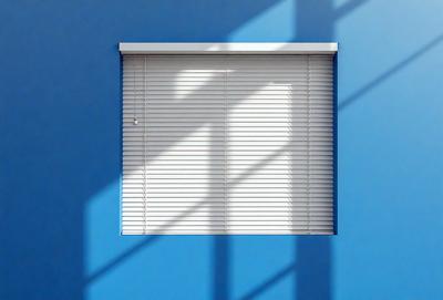 The primary subject of the image is a window with a white shade. The window is located on a blue wall, which serves as the setting or background. The visual style of the image is a photo, capturing the natural light shining through the window. The colors and mood of the image are predominantly blue, creating a calm and serene atmosphere.