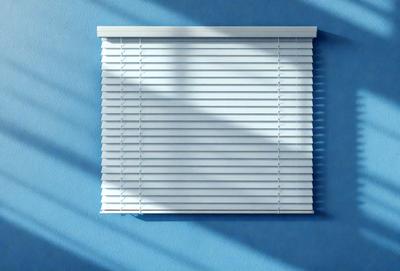 The primary subject of the image is a white shade, which is a part of a window blind. The shade is hanging on a blue wall, creating a contrast between the white shade and the blue background. The visual style of the image is a photo, capturing the details of the shade and the wall. The colors and mood of the image are predominantly blue, with the white shade adding a touch of brightness to the scene.