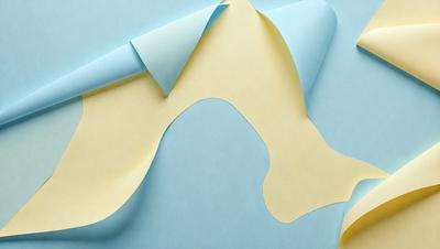 The primary subject of the image is a yellow paper cutout of a mountain. The setting is a blue surface, which serves as the background. The visual style is a combination of photo and digital art, as the paper cutout appears to be a photograph of a digital art piece. The colors and mood of the image are predominantly blue and yellow, creating a sense of depth and contrast.