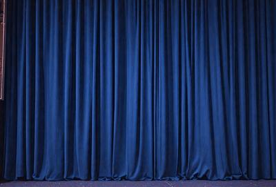 The primary subject of the image is a blue curtain, which is the main object, scene, or focus of the image. The setting is a stage or theater, with the curtain hanging down from the ceiling. The visual style is a photo, capturing the curtain in its natural environment. The colors and mood of the image are predominantly blue, creating a calm and serene atmosphere.