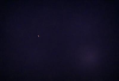 The primary subject of the image is a small, bright moon in the night sky. The setting is a dark, cloudy night with a purple hue. The visual style is a photo of the moon and the night sky. The colors and mood of the image are dark and moody, with the moon being the only bright spot in the sky.