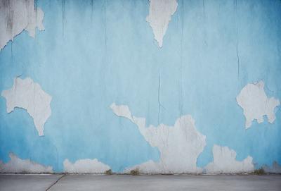 The primary subject of the image is a blue wall with white paint chipping off of it. The setting is an outdoor environment, with the wall being the main object of focus. The visual style is a photo, capturing the chipped paint and the blue wall. The colors and mood of the image are predominantly blue and white, with the white paint chipping off creating a sense of wear and tear.