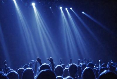 The primary subject of the image is a large group of people standing in a darkened auditorium, with their arms raised in the air. The setting is a concert or event, as evidenced by the stage lights illuminating the crowd. The visual style is a photo, capturing the excitement and energy of the event. The colors and mood are predominantly dark, with the stage lights providing a contrasting brightness to the scene.