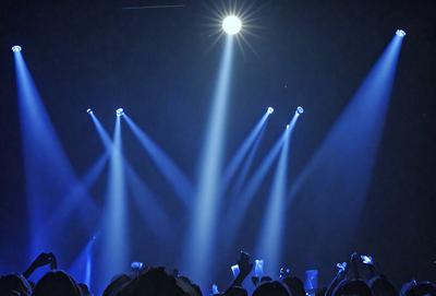 The primary subject of the image is a group of people standing in a dark room, with their arms raised. The setting is a concert or event, as indicated by the lights shining on the crowd. The visual style is a photo, capturing the moment of the people enjoying the event. The colors and mood are dark and energetic, as the crowd is engaged in the performance.