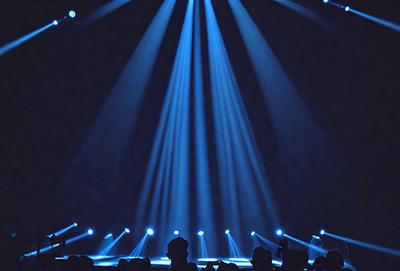 The primary subject of the image is a person standing in a darkened room, with a blue light illuminating the area. The setting appears to be a stage or a similar environment, as the person is surrounded by a crowd of people. The visual style of the image is a black and white photo, which adds a dramatic and artistic touch to the scene. The dominant colors in the image are blue and black, creating a moody and atmospheric ambiance.