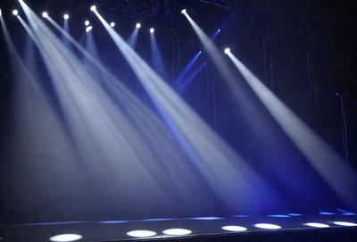 The primary subject of the image is a stage with a blue background. The stage is illuminated by a series of bright lights, creating a dramatic and captivating atmosphere. The lights are positioned at various angles and heights, creating a visually striking scene. The setting appears to be a theater or a similar venue, where performances or events take place. The visual style of the image is a combination of photography and digital art, with the lights and stage creating a dynamic and engaging composition. The colors and m