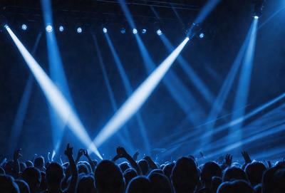 The primary subject of the image is a large crowd of people gathered in a darkened auditorium. The setting is a stage with a blue light illuminating the audience. The visual style is a black and white photo, which adds a dramatic and artistic touch to the scene. The colors and mood are predominantly dark and moody, creating a sense of excitement and anticipation for the event taking place.
