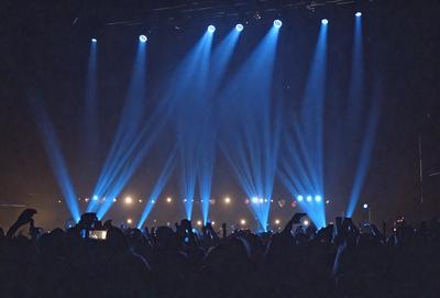 The primary subject of the image is a large crowd of people gathered in a darkened auditorium. The setting is a stage with a blue light illuminating the audience. The visual style is a photo, capturing the excitement and energy of the event. The colors and mood are predominantly dark, with the blue light creating a dramatic atmosphere.