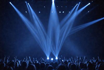 The primary subject of the image is a large crowd of people gathered in a darkened auditorium. The setting is a stage with bright lights illuminating the audience. The visual style is a photo, capturing the excitement and energy of the event. The colors and mood are predominantly dark, with the bright lights creating a contrasting effect.