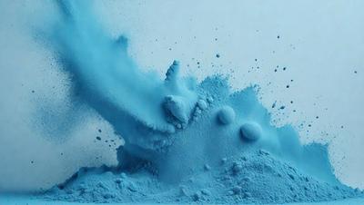The primary subject of the image is a blue substance, possibly a powder or a liquid, that is being blown or sprayed into the air. The setting appears to be a white background, which contrasts with the blue substance. The visual style of the image is a close-up of the blue substance, showcasing its texture and movement. The colors and mood of the image are predominantly blue, creating a sense of movement and dynamism.