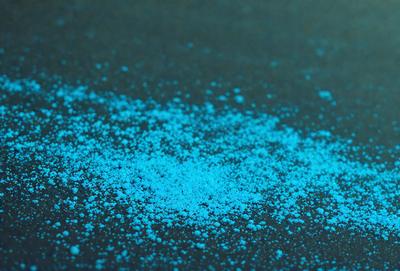 The primary subject of the image is a blue powder or substance, which is spread across a black surface. The setting appears to be a dark room, and the visual style is a close-up of the blue powder. The dominant colors in the image are blue and black, creating a moody and dramatic atmosphere.