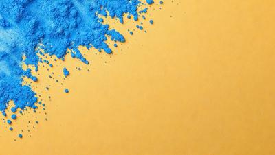 The primary subject of the image is a blue substance, possibly a powder or a liquid, that is spreading or dripping onto a yellow surface. The setting appears to be a table or a countertop, with the blue substance creating a striking contrast against the yellow background. The visual style of the image is a mix of digital art and illustration, as the blue substance is depicted in a somewhat abstract manner. The colors and mood of the image are vibrant and energetic, with the blue substance