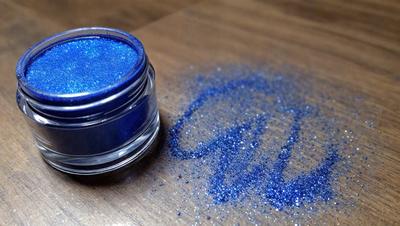 The primary subject of the image is a bottle of blue glitter nail polish. The setting is a wooden table, and the visual style is a close-up of the bottle. The dominant colors are blue and silver, creating a sparkly and shiny appearance. The overall atmosphere is vibrant and eye-catching, with the glittery blue nail polish as the main focus.