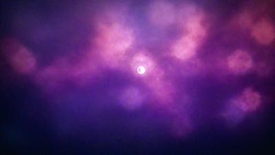 The primary subject of the image is a sun, which is shining brightly through a purple sky. The setting is a cloudy sky, and the visual style appears to be a photo. The dominant colors in the image are purple and orange, creating a moody and dramatic atmosphere.