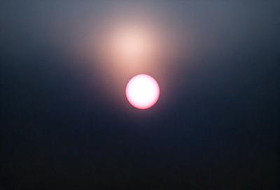 The primary subject of the image is a bright, pink sun rising above a dark, cloudy sky. The sun is positioned in the upper-middle part of the scene, with its rays extending outwards. The setting is a nighttime sky, with the sun being the only source of light. The visual style of the image is a photo, capturing the natural beauty of the sunrise. The colors and mood of the image are dominated by the contrasting colors of the pink sun and the dark, cloud