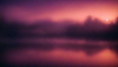 The primary subject of the image is a body of water, which appears to be a lake. The setting is a foggy, misty evening, creating a serene and atmospheric scene. The visual style is a photo, capturing the beauty of the lake and its surroundings. The colors and mood of the image are predominantly purple and pink, giving it a warm and dreamy ambiance.